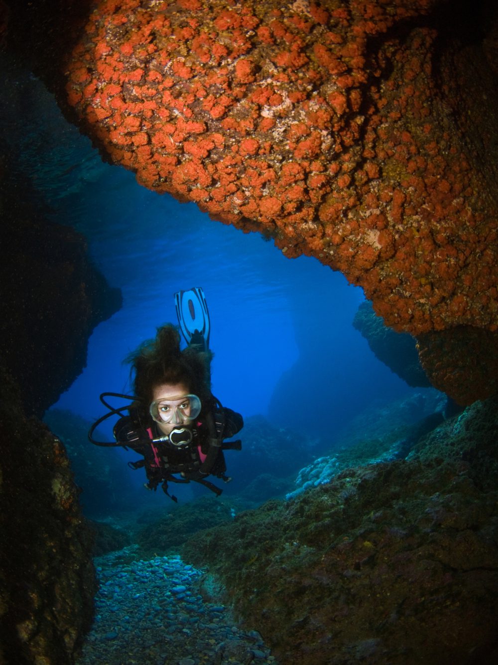 Young girl doing scuba-diving and exploring an underwater cave with walls covered by orange coral.